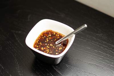 Mørk, glinsende sød chilisauce med tørrede chiliflager i hvid firkantet skål og med sølvfarvet ske