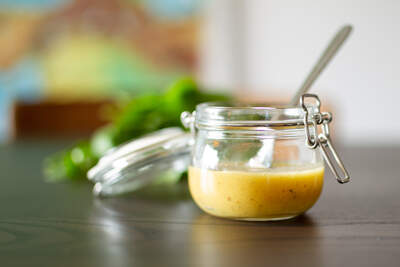 Gylden vinaigrette i glaskrukke med tæt, cremet tekstur og grøn pynt i baggrunden.