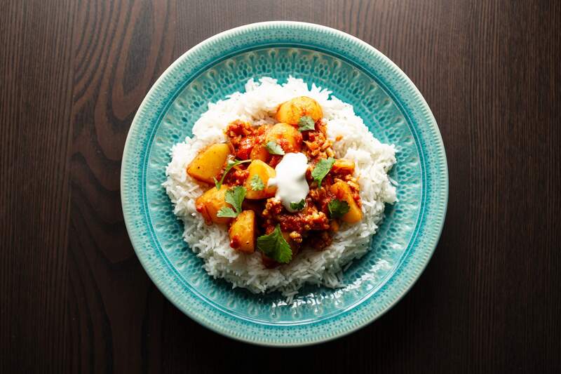 Aloo Gobi-ret med kartofler og blomkål i rød-orange sauce, hvid ris, frisk koriander og yoghurt på en turkis tallerken.