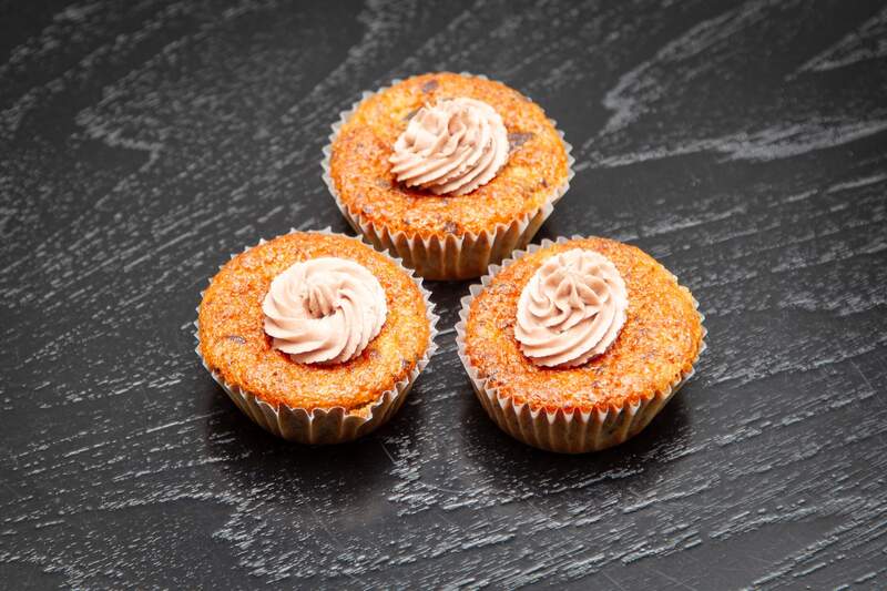 Tre bananmuffins med lysebeige frosting, sprøde kanter og saftig, gylden midte på sort bord.