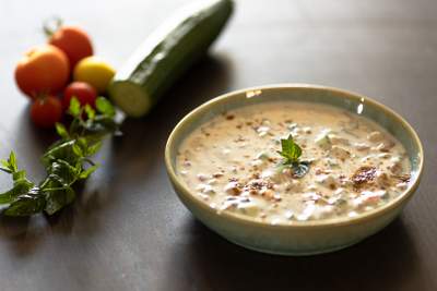 Skål med cremet raita drysset med spidskommen og pyntet med frisk mynte, med agurk, tomater og mynte i baggrunden