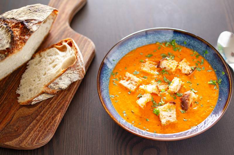 Krydret peberfrugtsuppe i blå skål med sprøde parmesan-croutoner, pyntet med persille; ved siden af rustikt brød.