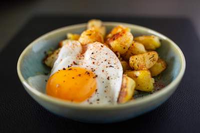 Brasede kartofler og spejlæg i en blå skål; gyldne kartoffelstykker, blank æggehvide og rød paprika.