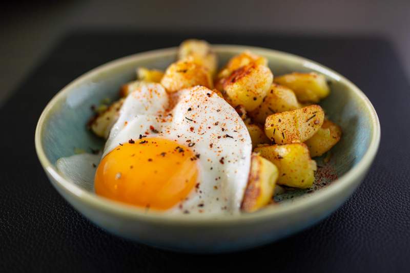 Brasede kartofler og spejlæg i en blå skål; gyldne kartoffelstykker, blank æggehvide og rød paprika.