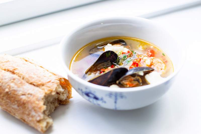 Fiskesuppe med muslinger i gylden bouillon, cremet konsistens, hvide fiskestykker, røde peberfrugter og sprø brød ved siden.