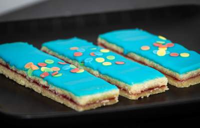 Tre hindbærsnitter med blå glasur, sprøde kanter, farverige tivolidrys og syltetøjslag.