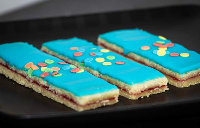 Tre hindbærsnitter med blå glasur, sprøde kanter, farverige tivolidrys og syltetøjslag.