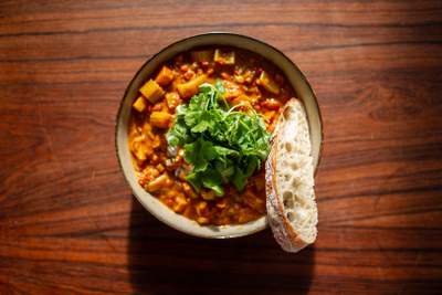 En dyb skål med orange mulligatawny-suppe, terninger af gulerødder og kartofler, frisk koriander og brød ved siden.