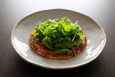 Gyldenbrunt sprødt fladbrød dækket af friske, mørkegrønne ruccola på en hvid tallerken.